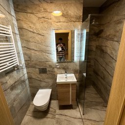 REMBUD - Nowoczesna łazienka z marmurowymi płytkami, kabiną prysznicową ze szklaną ścianką, podświetlanym lustrem, umywalką z szafką i grzejnikiem drabinkowym. W lustrze odbija się osoba robiąca zdjęcie.