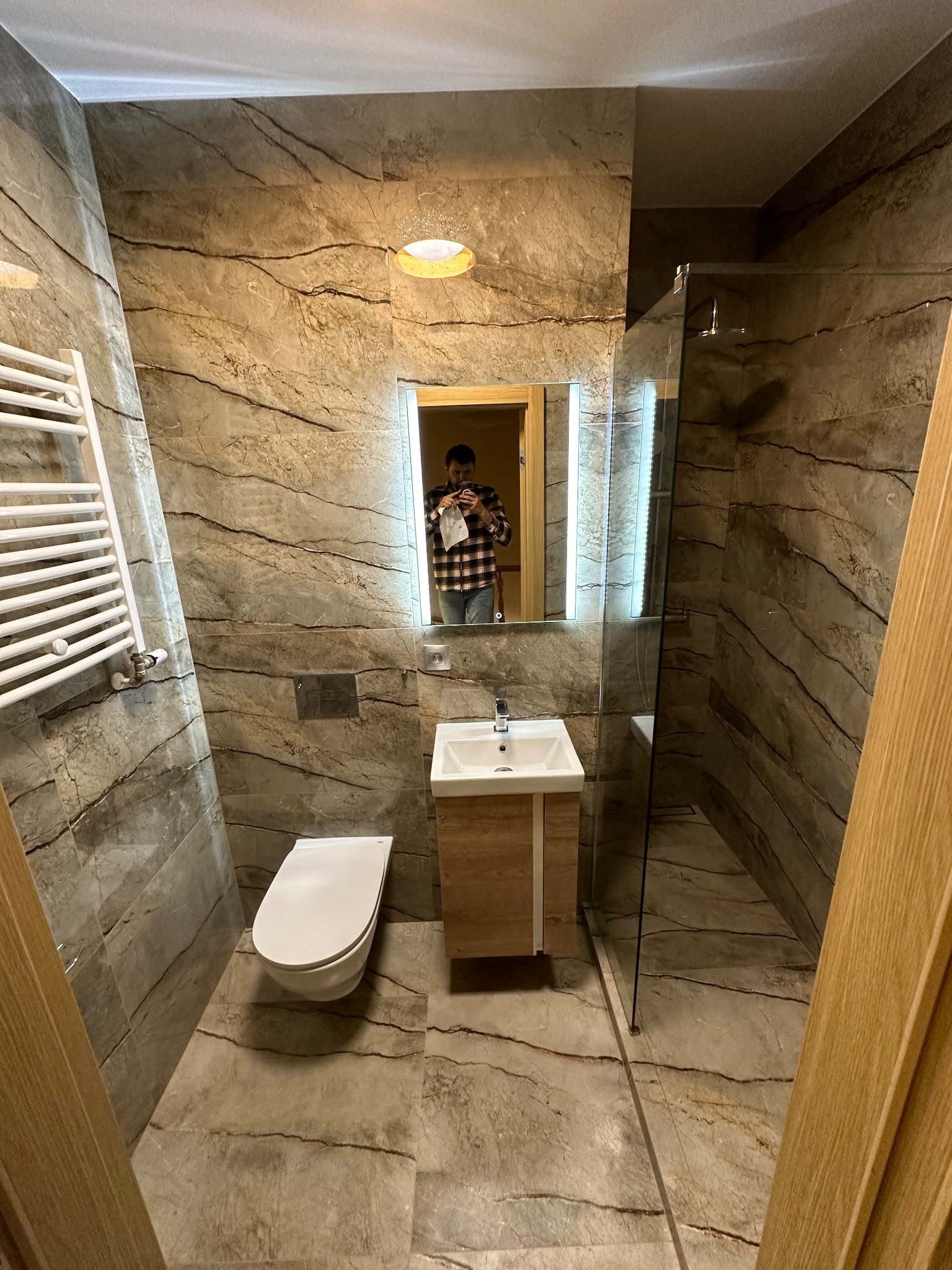 Nowoczesna łazienka z marmurowymi płytkami, kabiną prysznicową ze szklaną ścianką, podświetlanym lustrem, umywalką z szafką i grzejnikiem drabinkowym. W lustrze odbija się osoba robiąca zdjęcie.