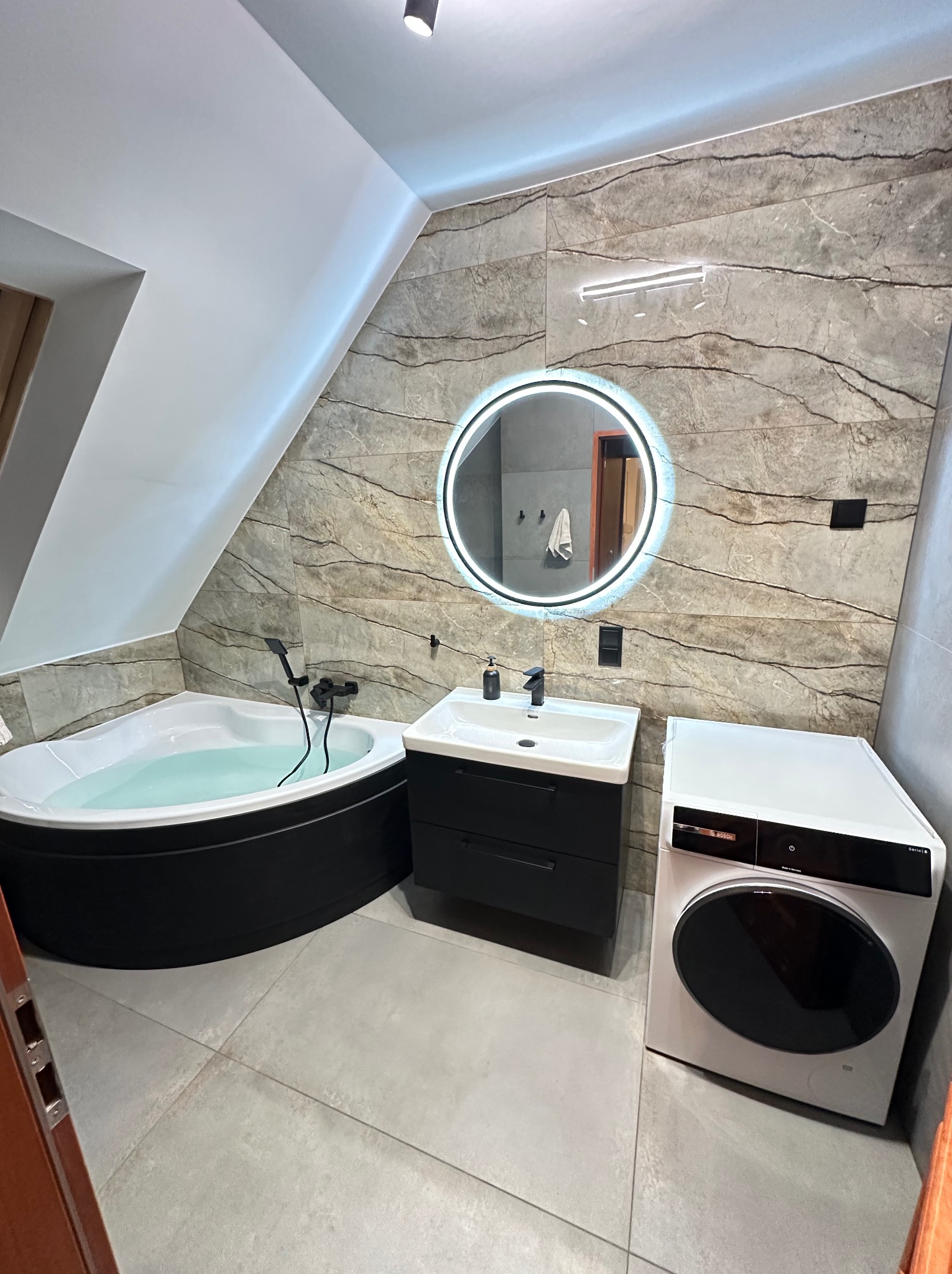 Nowoczesna łazienka na poddaszu z wanną wypełnioną wodą, okrągłym lustrem z podświetleniem LED, czarną szafką pod umywalką i pralką Bosch Serie 8.