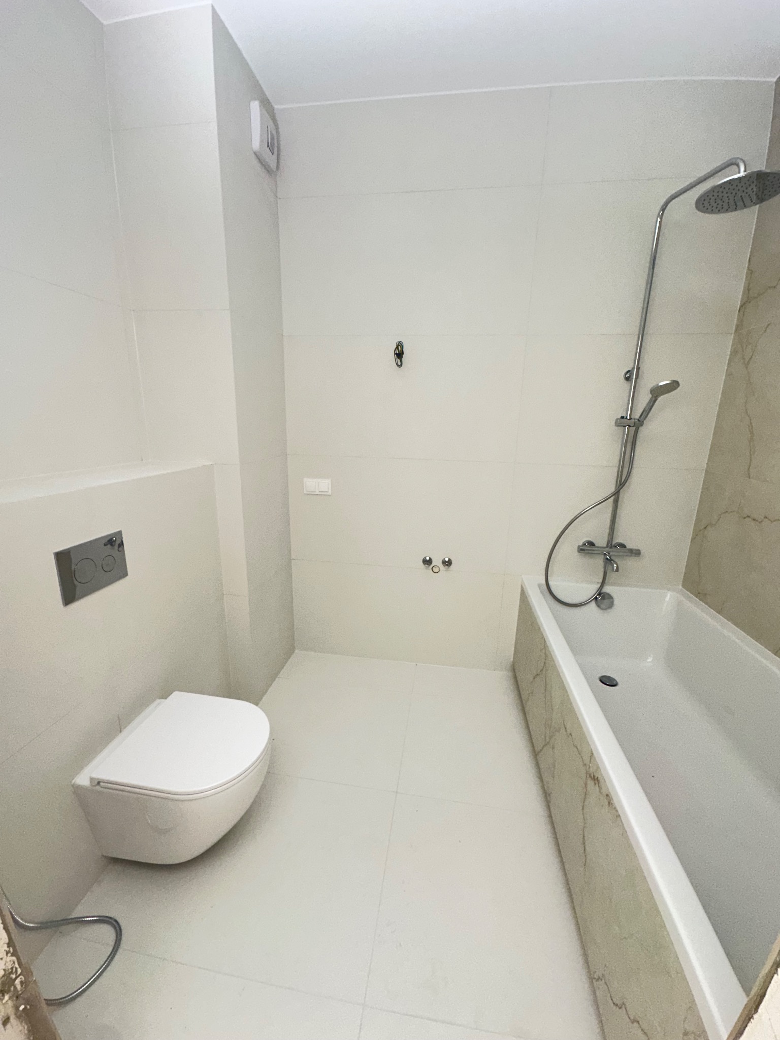 Jasna łazienka po remoncie: białe płytki, podwieszana toaleta, wanna z obudową imitującą marmur, chromowana armatura. Minimalistyczny design, czystość i nowoczesność.