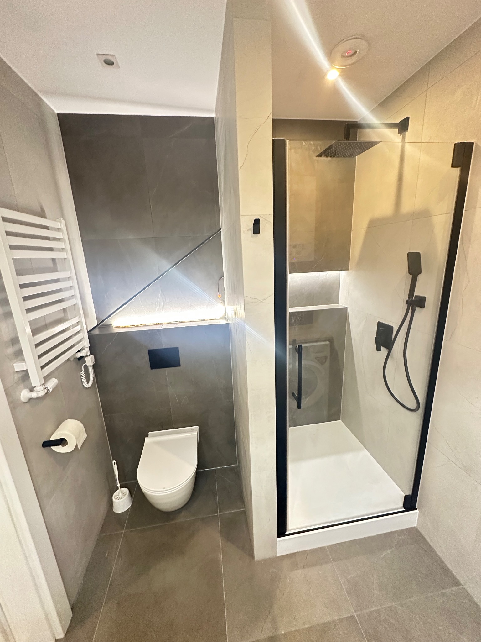 Nowoczesna łazienka z kabiną prysznicową typu walk-in z czarnymi ramami, podświetlaną półką i grzejnikiem drabinkowym. Minimalistyczny design, szaro-biała kolorystyka.