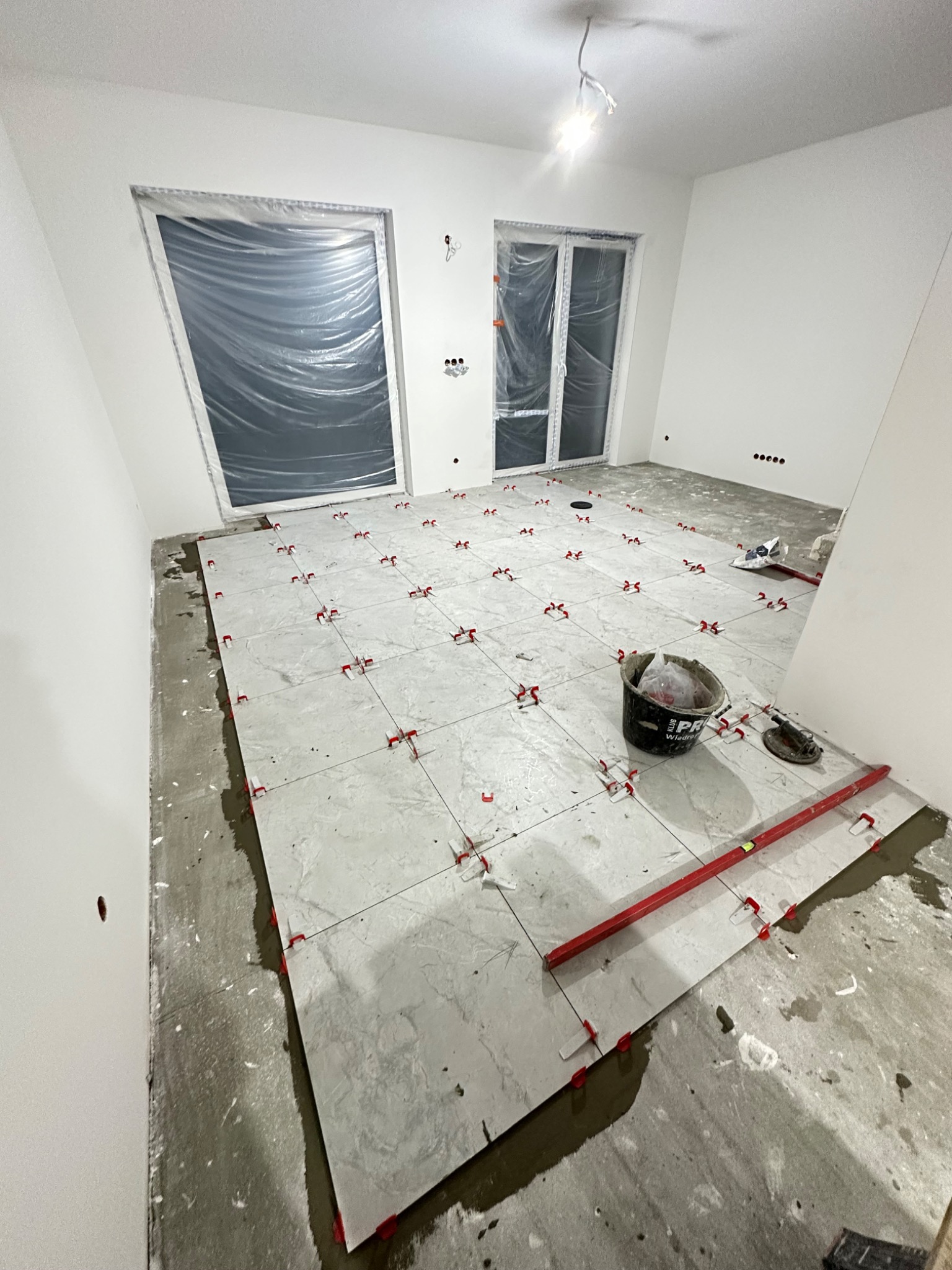 Układanie jasnych, marmurkowych płytek podłogowych w nowym, pustym pomieszczeniu. Widoczne poziomica, wiadro i system poziomowania płytek.