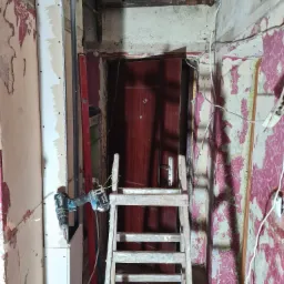 Montaż płyty karton gips przy drzwiach wejściowych, widoczna drabina, wiertarka akumulatorowa i fragment starej ściany z resztkami tapety.