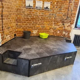 Podwyższona platforma z ciemnych paneli podłogowych, z dwustopniowym wejściem, ustawiona przy ceglanej ścianie z plakatami miast, z widocznym wiadrem i kuwetą malarską.