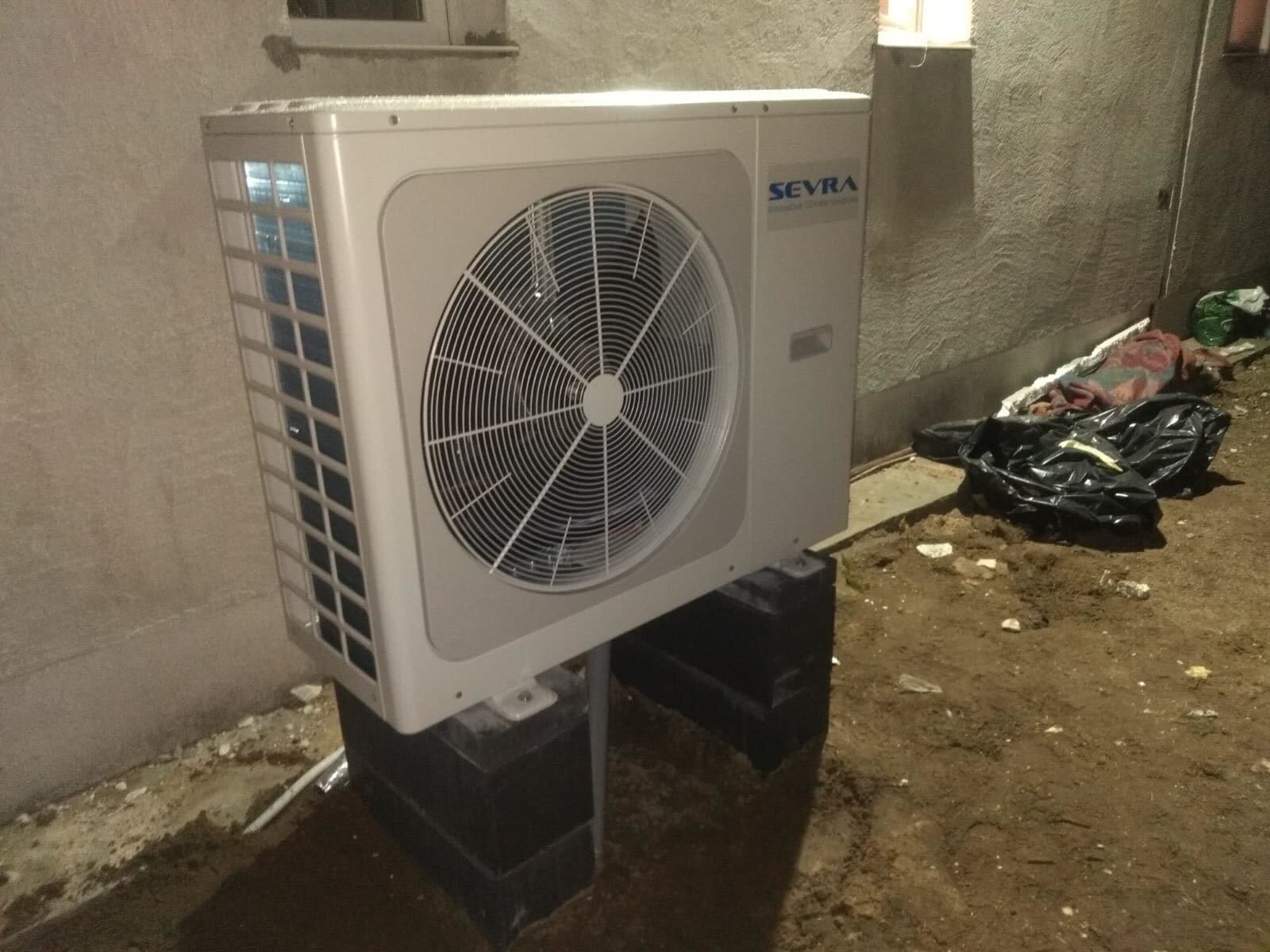 Biała zewnętrzna jednostka pompy ciepła marki Sevra, zamontowana na czarnych blokach betonowych przy ścianie budynku, z ziemią i śmieciami w tle.