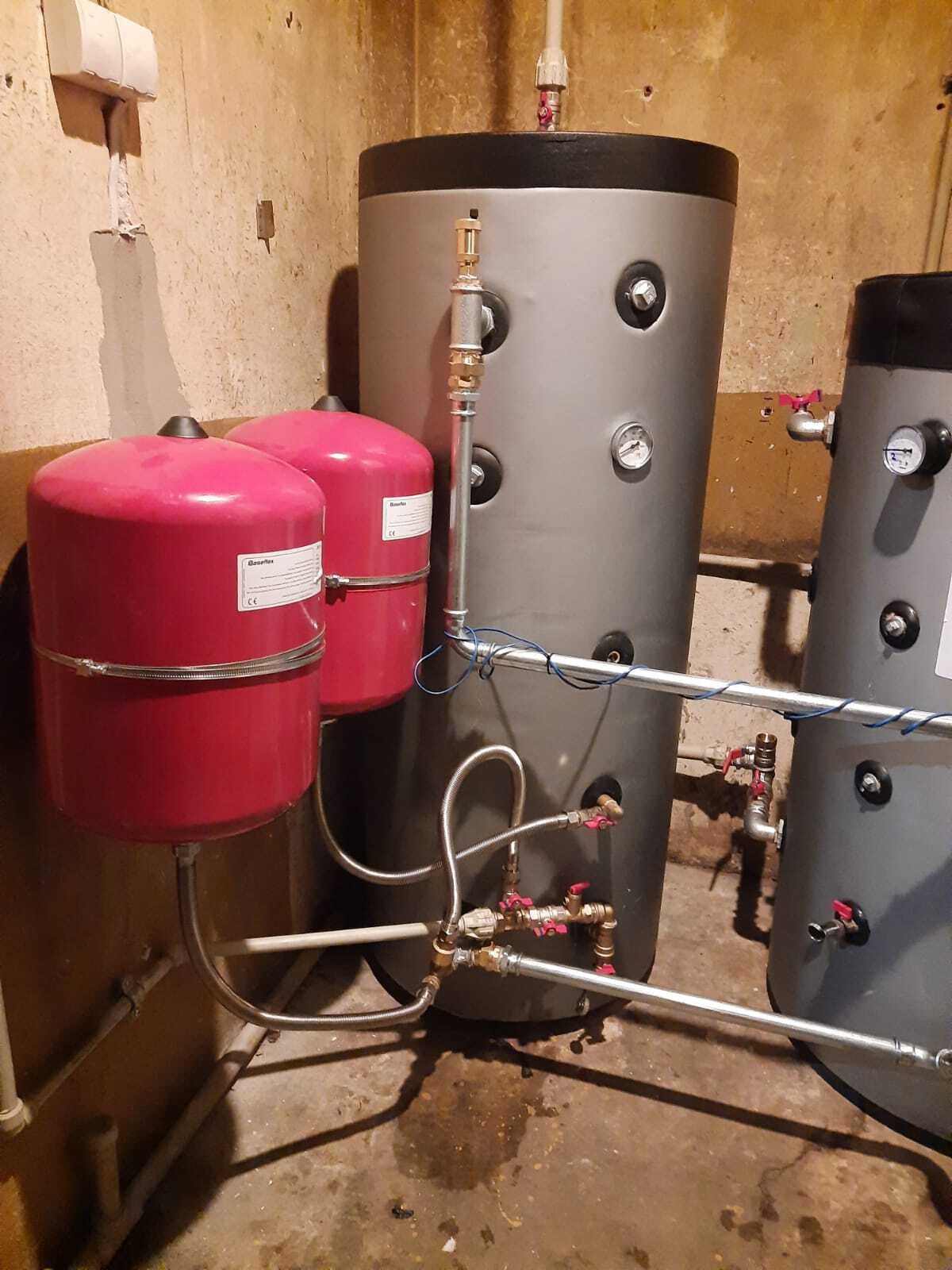 Dwa czerwone zbiorniki wyrównawcze obok szarych akumulatorów ciepła z podłączonymi rurami i zaworami w pomieszczeniu o surowych ścianach.