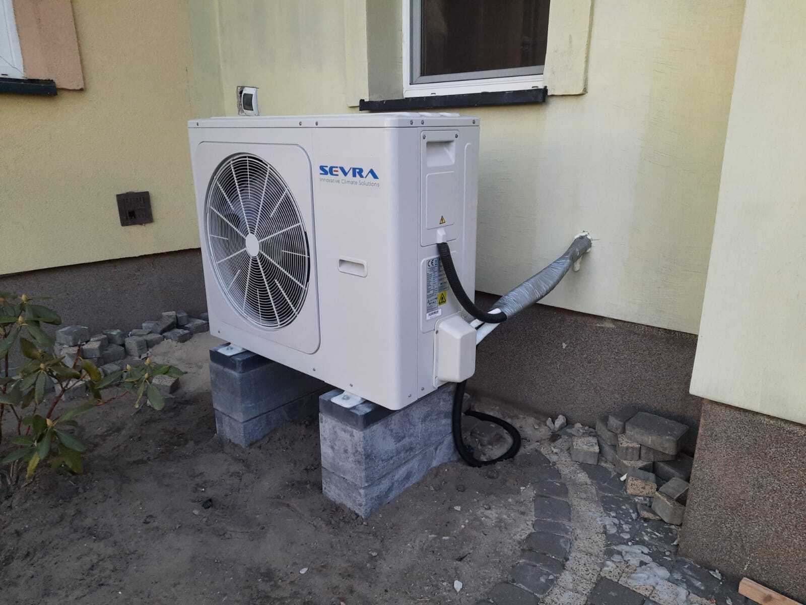 Zewnętrzna jednostka pompy ciepła marki Sevra, zamontowana na betonowych bloczkach przy ścianie budynku, z widocznymi przewodami w izolacji.