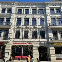 Fasada odnowionej kamienicy z elementami dekoracyjnymi, balkony z kutymi balustradami, parter z lokalami usługowymi, widoczni przechodnie.