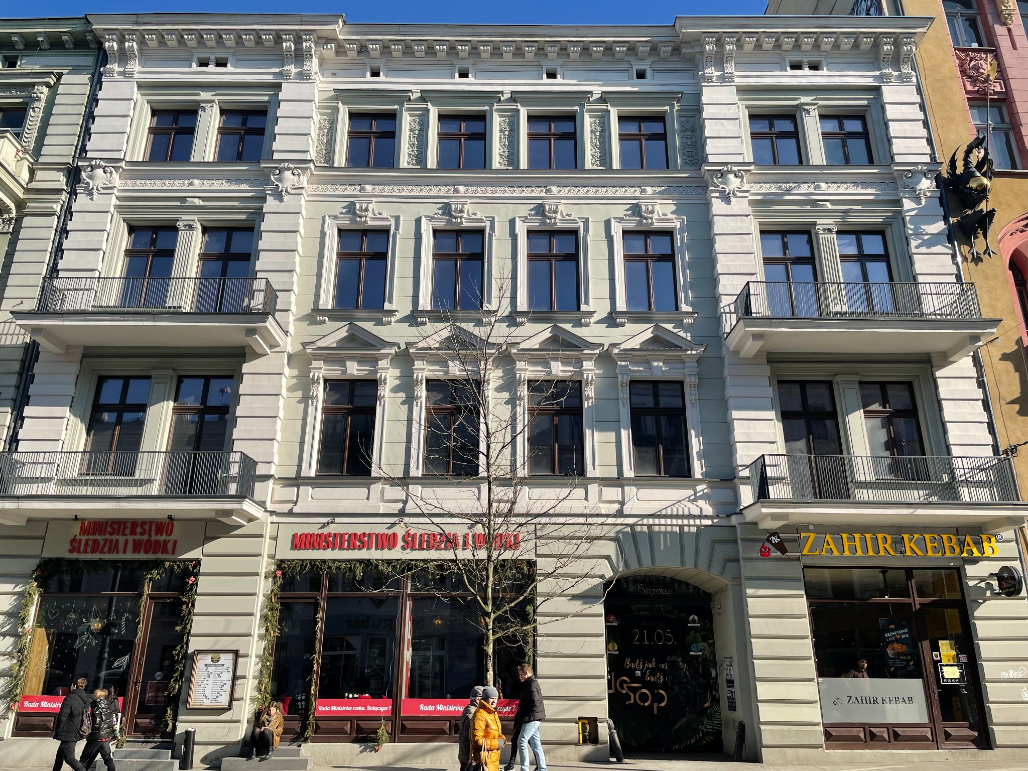 Fasada odnowionej kamienicy z elementami dekoracyjnymi, balkony z kutymi balustradami, parter z lokalami usługowymi, widoczni przechodnie.