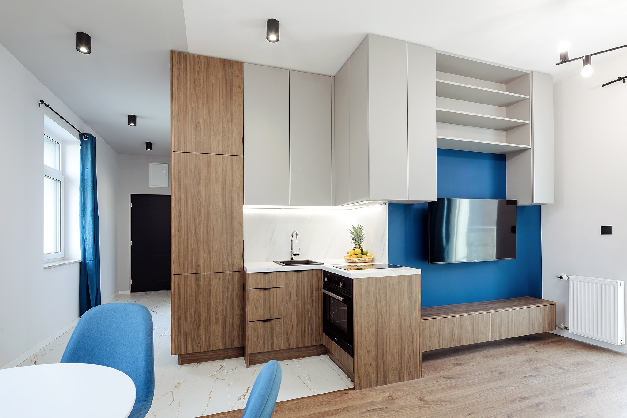 Nowoczesna kuchnia z drewnianymi i białymi szafkami, marmurowym blatem, niebieską ścianą z telewizorem, drewnianą podłogą i błękitnymi krzesłami w tle.