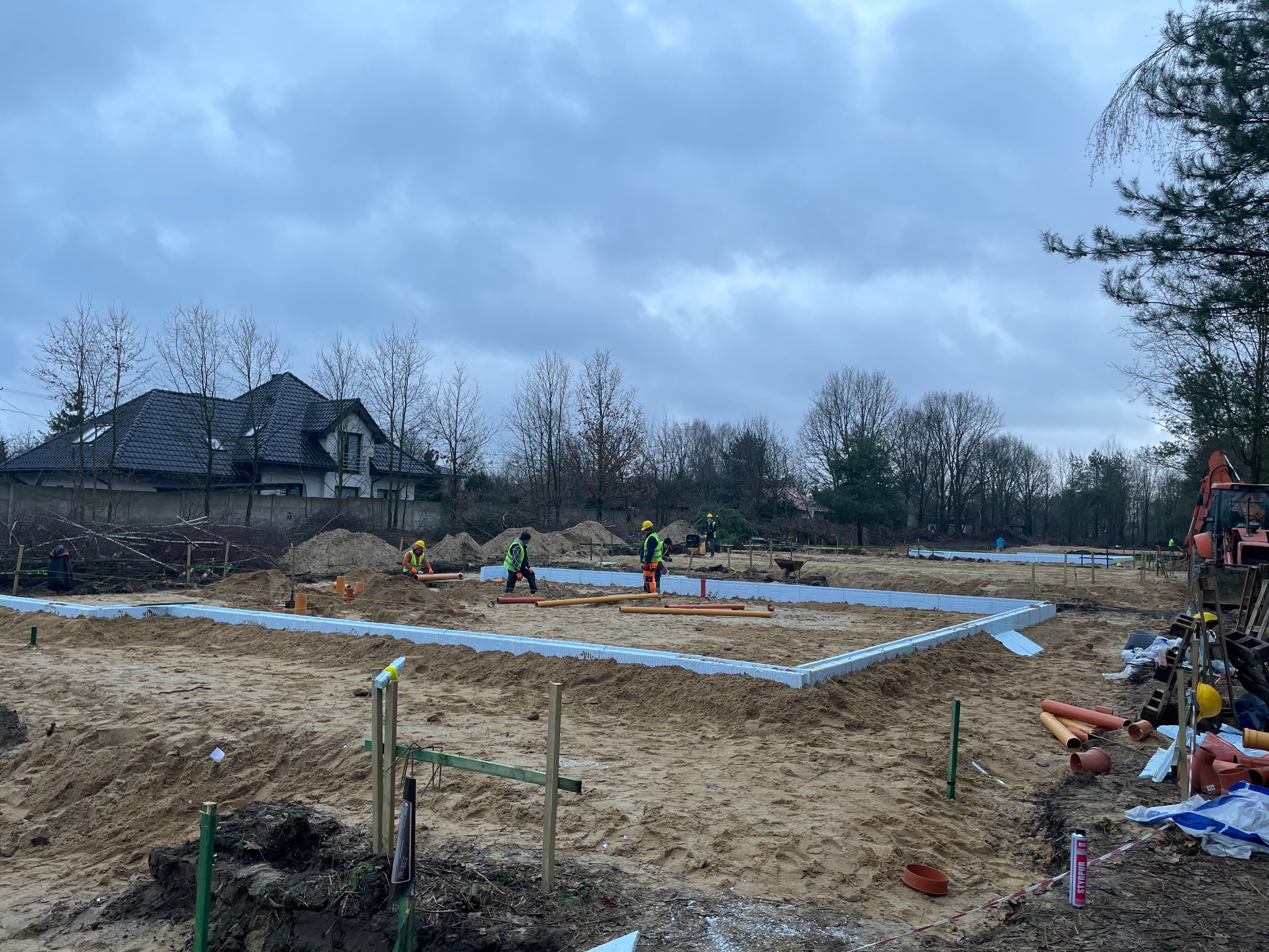 Budowa fundamentów domu jednorodzinnego: ekipa w odblaskowych kamizelkach układa izolację termiczną w wykopie, widoczne rury kanalizacyjne i koparka w tle.