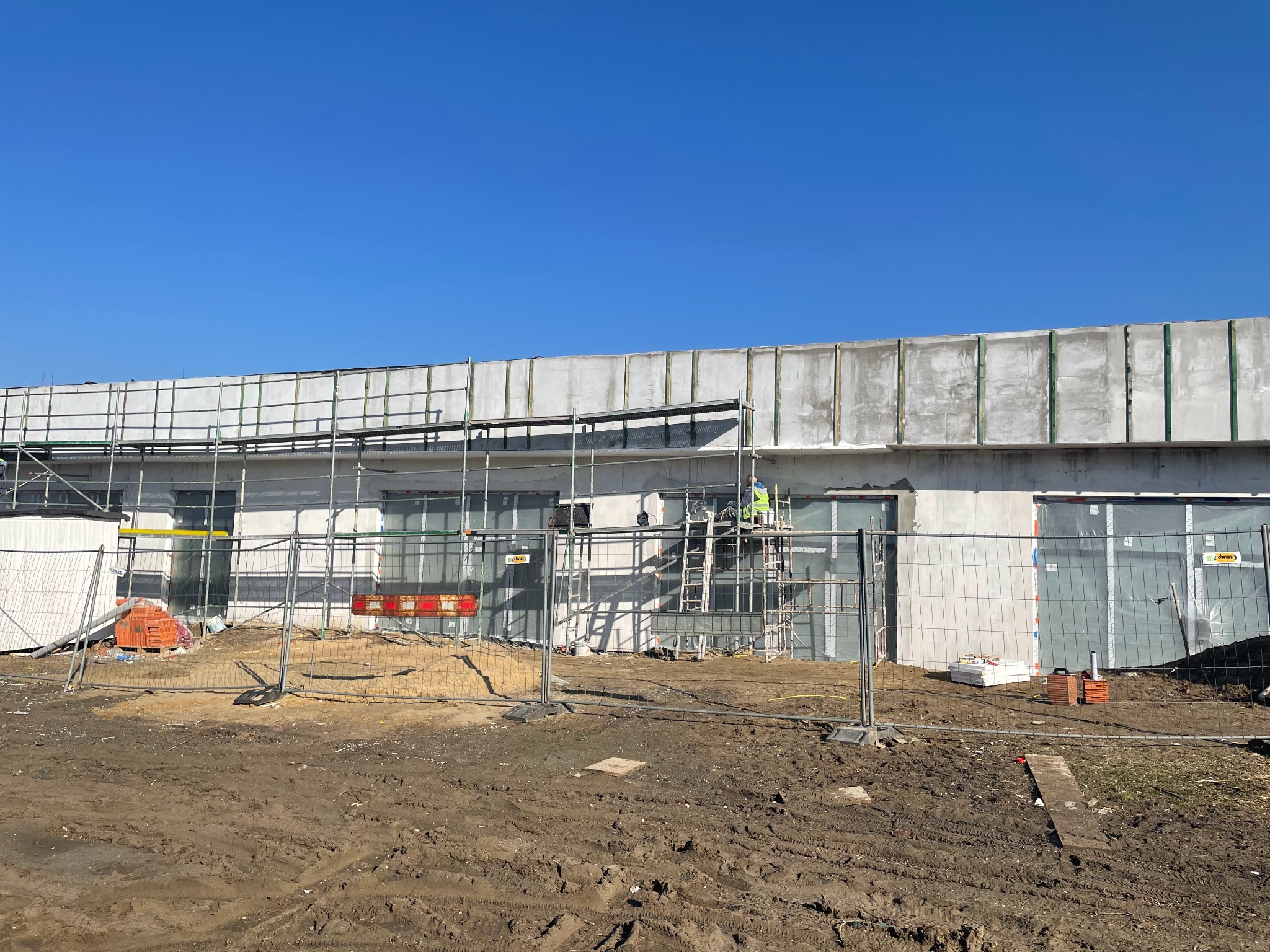 Budynek w trakcie ocieplania elewacji, widoczne rusztowania, siatka zabezpieczająca i pracownik na drabinie; teren budowy z błotnistym podłożem.