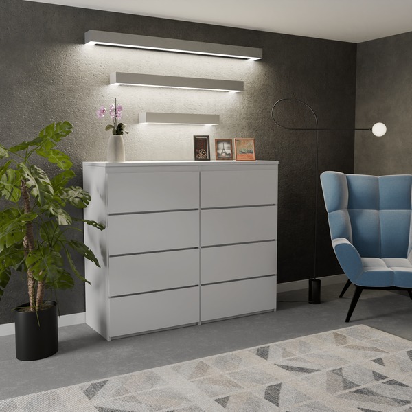 Biała komoda z ośmioma szufladami bez uchwytów, ustawiona w pokoju z szarą ścianą, oświetlona trzema podłużnymi lampami LED, z rośliną doniczkową po lewej stronie i błękitnym fotelem w tle.