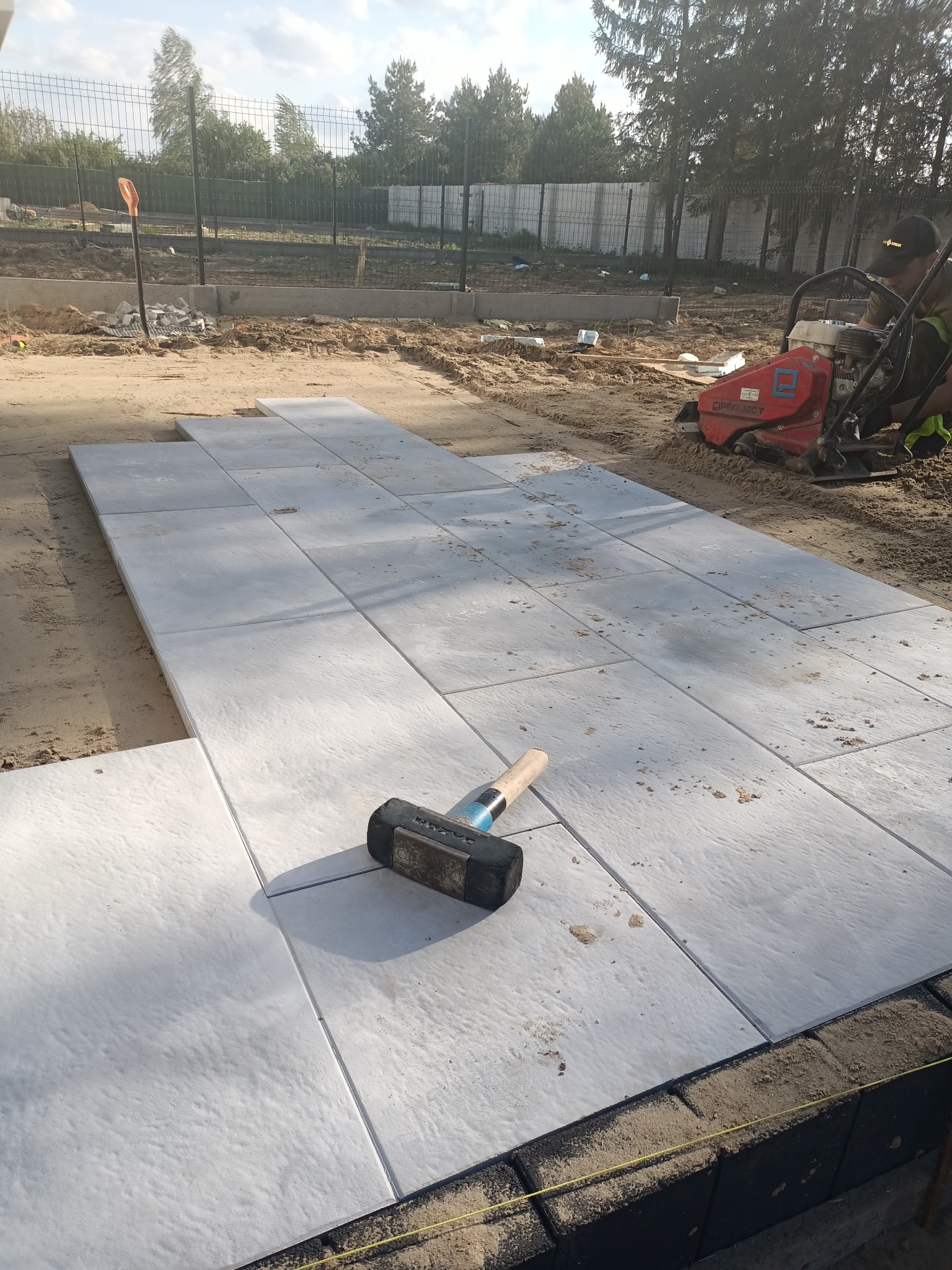 Układanie jasnoszarej kostki brukowej na podsypce piaskowej, widoczny gumowy młotek, ubijarka spalinowa i przygotowane podłoże pod obrzeża.
