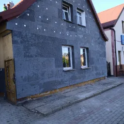 Dom w trakcie ocieplania: szara elewacja pokryta płytami izolacyjnymi z widocznymi białymi kołkami mocującymi, dwie białe okna na parterze i dwa na poddaszu, pracownik na dachu przy kominie...