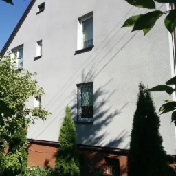 Jasnoszary dom jednorodzinny z brązowym dachem i podmurówką, widok z ogrodu z zielonymi drzewami iglastymi i liściastymi.