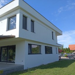 Józo - Nowoczesny, dwupiętrowy dom o minimalistycznej architekturze z gładką, białą elewacją i ciemnymi ramami okiennymi. Na parterze duże przesuwne drzwi tarasowe, obok zielony trawnik.