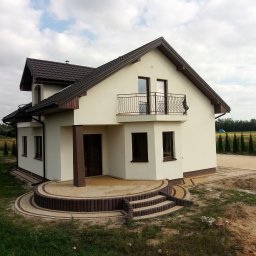 Józo - Nowo wykończony dom jednorodzinny z jasną elewacją, brązowym dachem i ozdobną, falistą balustradą balkonu. Przed wejściem okrągły taras z dwukolorowej kostki brukowej i schodami. W tle widoczny...