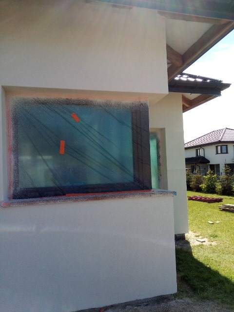 Fragment elewacji domu jednorodzinnego w trakcie prac wykończeniowych; okno zabezpieczone folią malarską, świeżo nałożony tynk, widoczne ślady farby w sprayu.