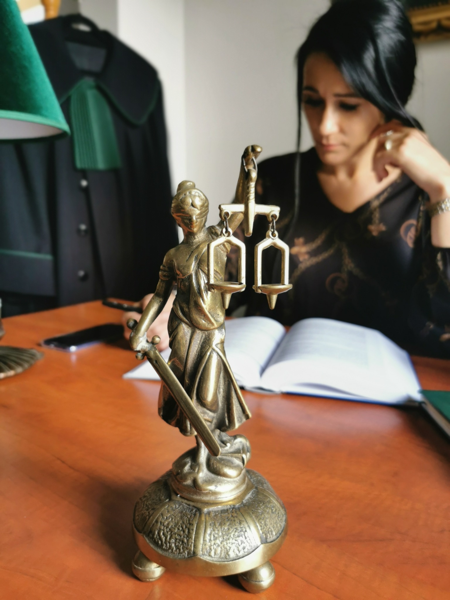 Statuetka Temidy na drewnianym biurku, w tle kobieta w czarnej sukience przegląda otwartą książkę, a na wieszaku wisi toga.