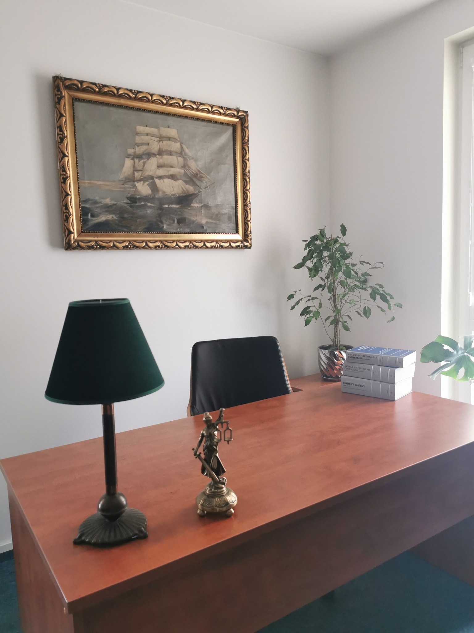 Gabinet prawny: biurko z drewnianym blatem, lampka z zielonym abażurem, statuetka Temidy, książki prawnicze i obraz z żaglowcem w złotej ramie.