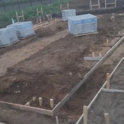 Przygotowanie fundamentów pod budowę domu: drewniane szalunki, wykopane rowy i ułożone bloczki betonowe Betard na placu budowy.