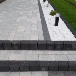 Schody i chodnik z szarej kostki brukowej, obrzeża z ciemnej kostki, białe kamienie dekoracyjne, trawnik w tle.