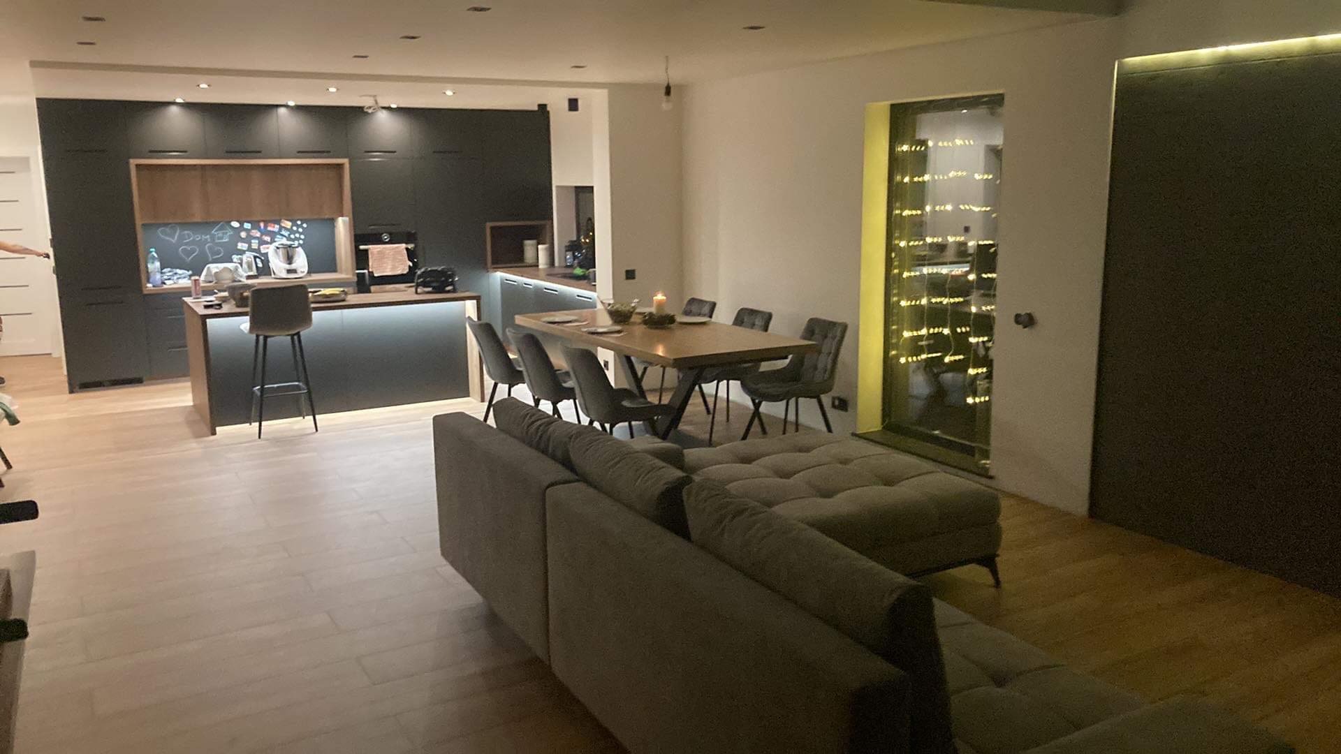 Nowoczesne wnętrze: parkiet z jasnego drewna, szara sofa, stół z krzesłami, czarna zabudowa kuchenna z drewnianymi elementami i wyspą, dekoracyjna ściana z oknem i lampkami.