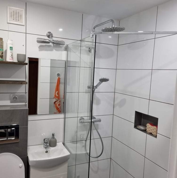 Nowoczesna łazienka z kabiną prysznicową walk-in, białe płytki kwadratowe z ciemną fugą, umywalka nablatowa na szafce, lustro z pomarańczowym ręcznikiem, półki z kosmetykami i wnęka z akcesoriami.