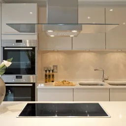 Elegancka, beżowa kuchnia z wyspą, na kt&oacute;rej stoi wazon z białymi kaliami, oświetlona punktowymi lampami podszafkowymi i srebrnym okapem.