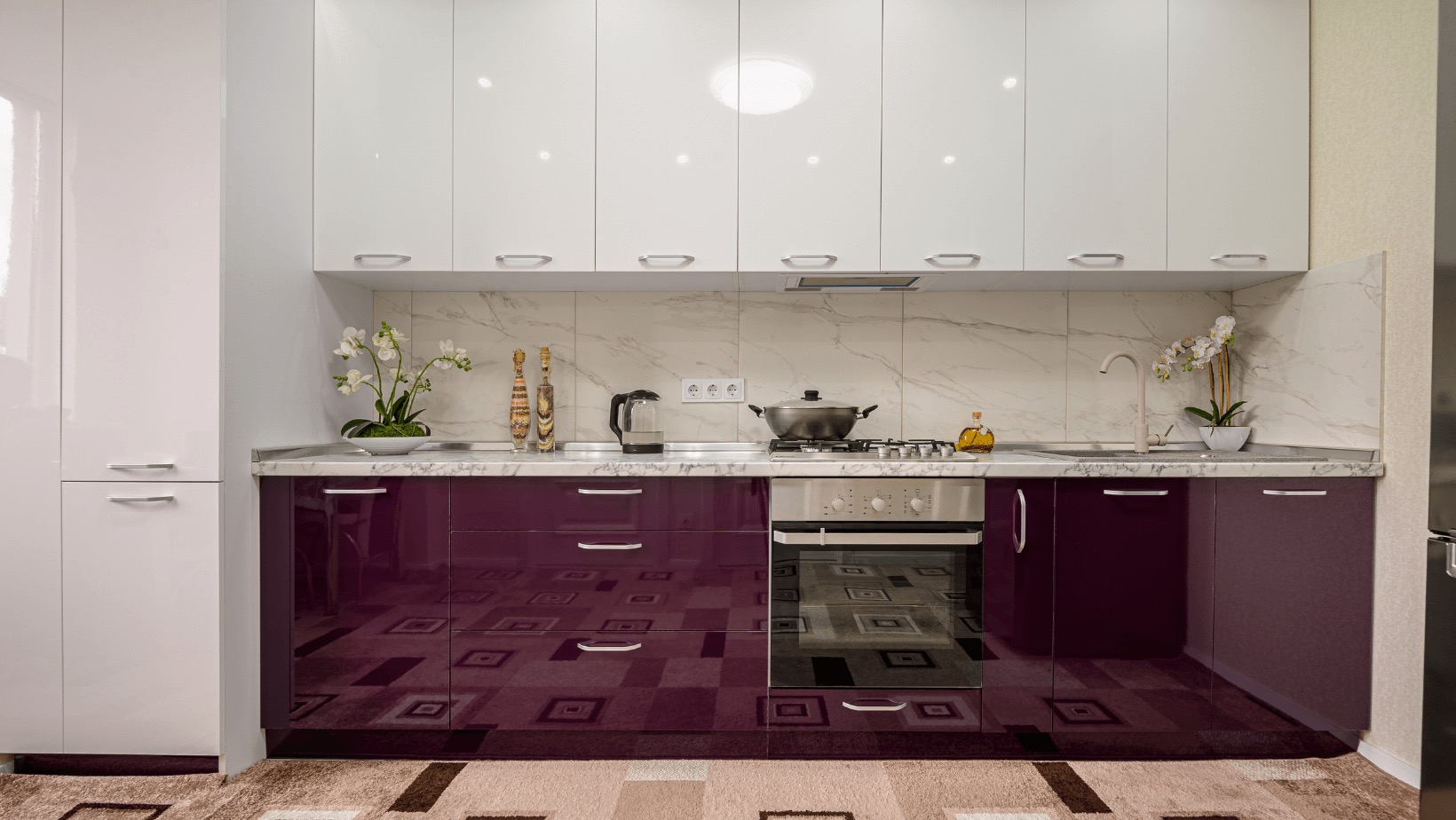 Nowoczesna kuchnia z białymi szafkami górnymi i bordowymi dolnymi, marmurowy backsplash, roślina w doniczce i akcesoria na blacie.