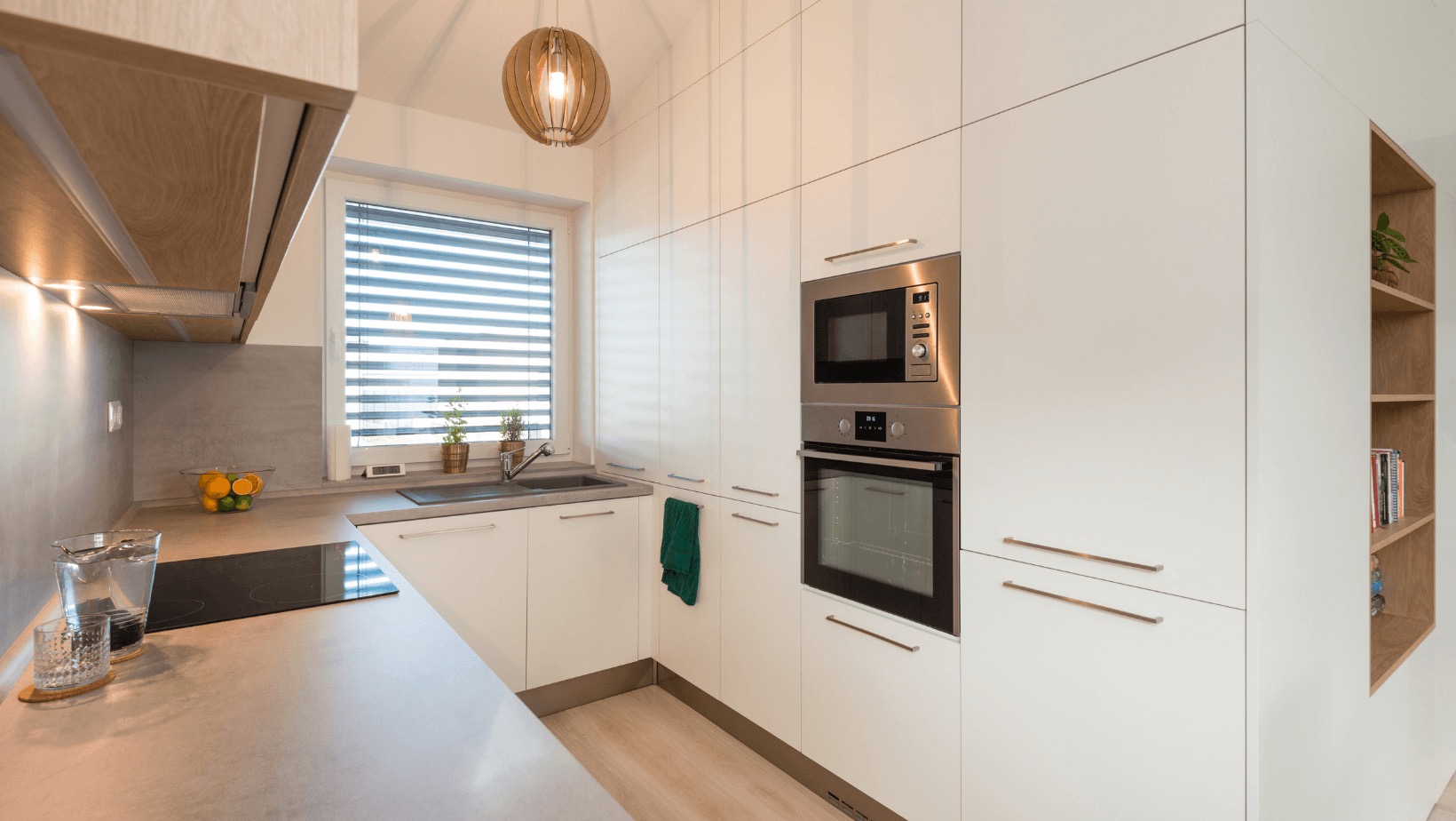 Nowoczesna, jasna kuchnia z białymi szafkami bez uchwytów, szarym blatem, zabudowanym piekarnikiem i mikrofalówką oraz drewnianą lampą wiszącą.