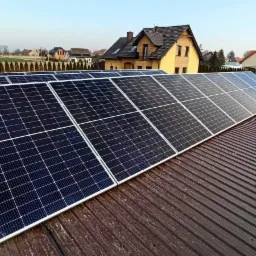 Panele fotowoltaiczne na brązowym dachu z blachy trapezowej, w tle domy jednorodzinne i pole uprawne.