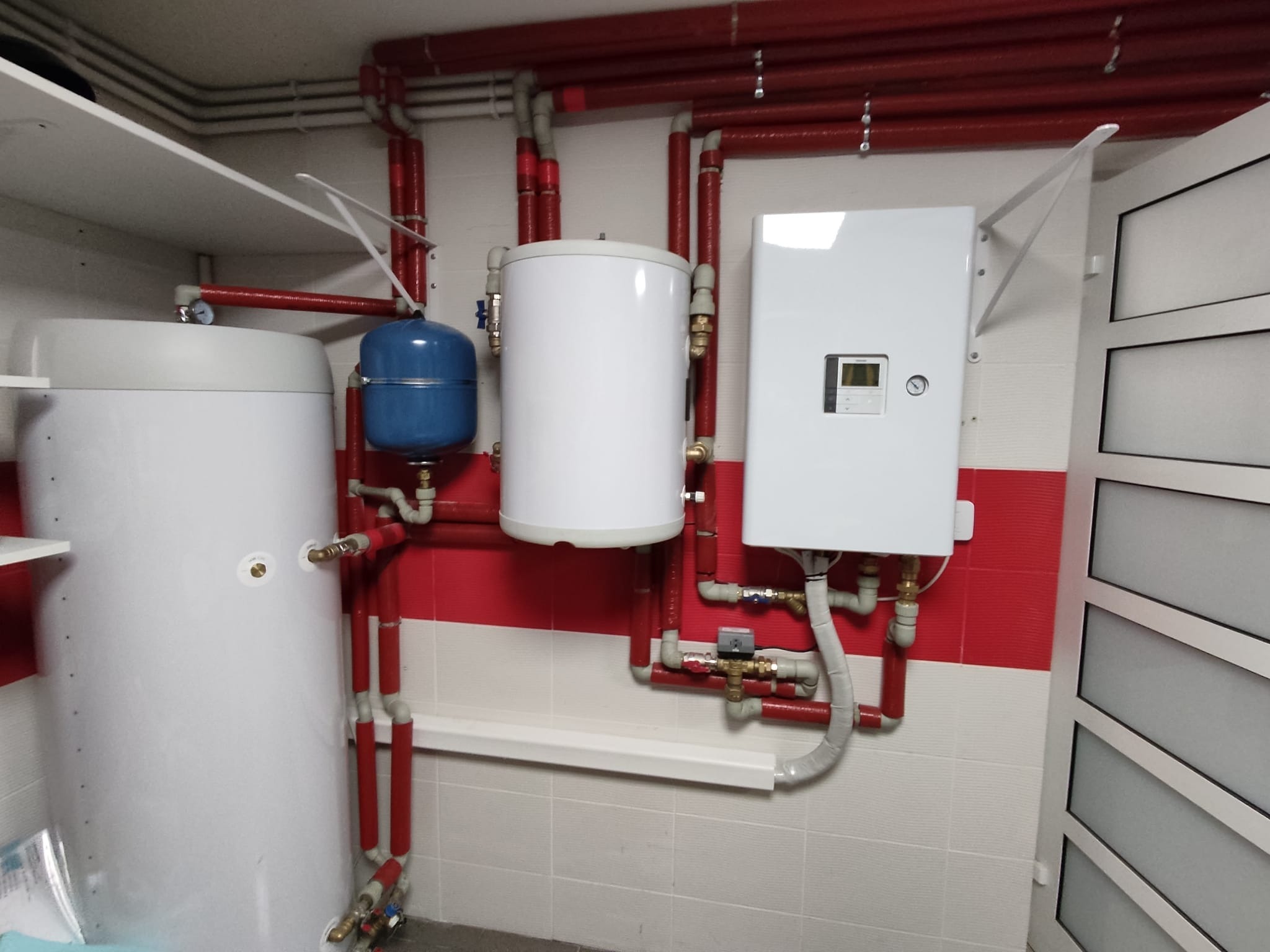Instalacja z dwoma białymi zbiornikami, niebieskim naczyniem wzbiorczym i białym panelem sterującym, z czerwonymi rurami, zamontowana w pomieszczeniu w Ostrowie Wielkopolskim, z płytkami...