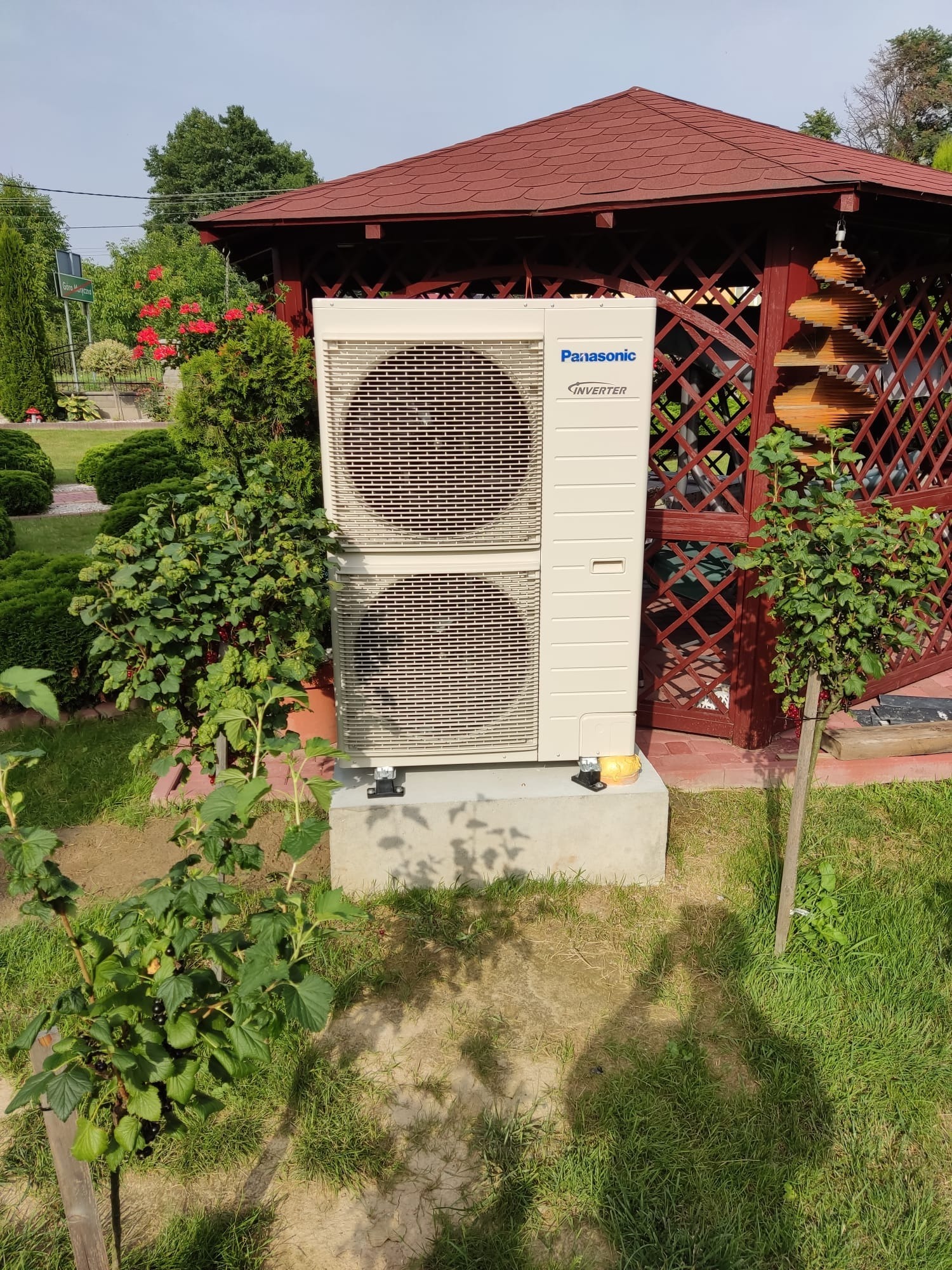 Zewnętrzna jednostka klimatyzacji Panasonic Inverter na betonowym fundamencie w ogrodzie, z czerwonym altanką w tle i krzewami wokół.