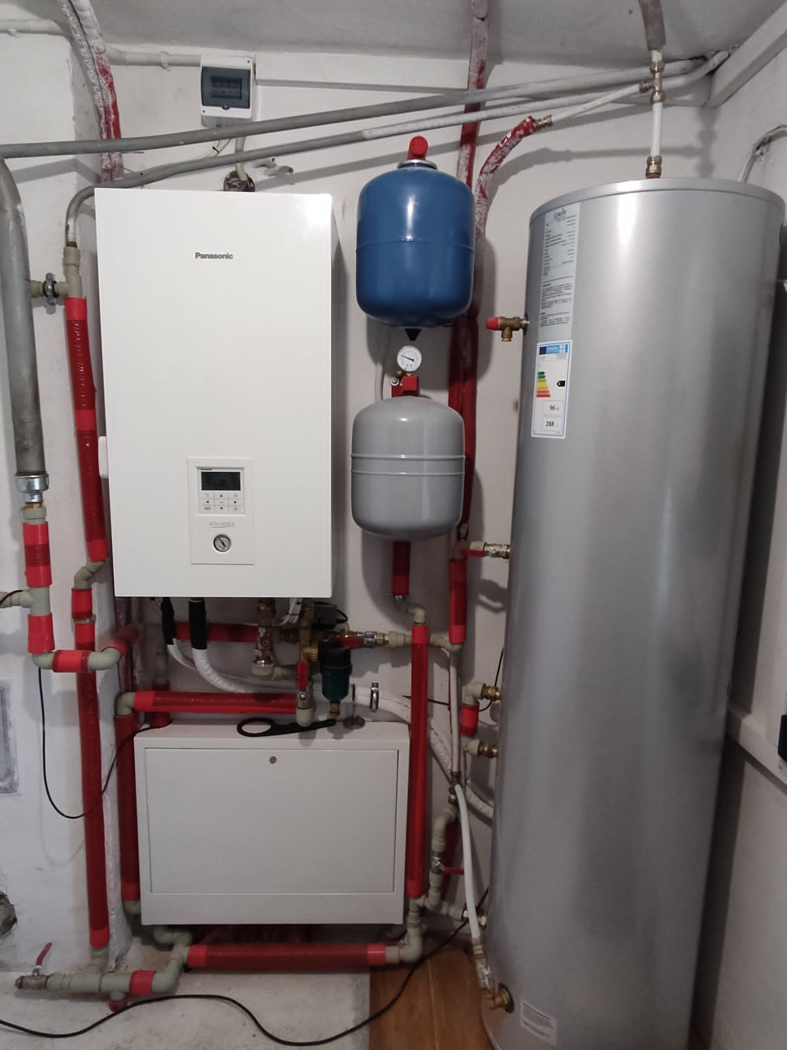 Instalacja grzewcza w piwnicy: biały piec Panasonic, dwa zbiorniki wyrównawcze (niebieski i szary), srebrny zasobnik ciepłej wody, czerwone rury instalacyjne.