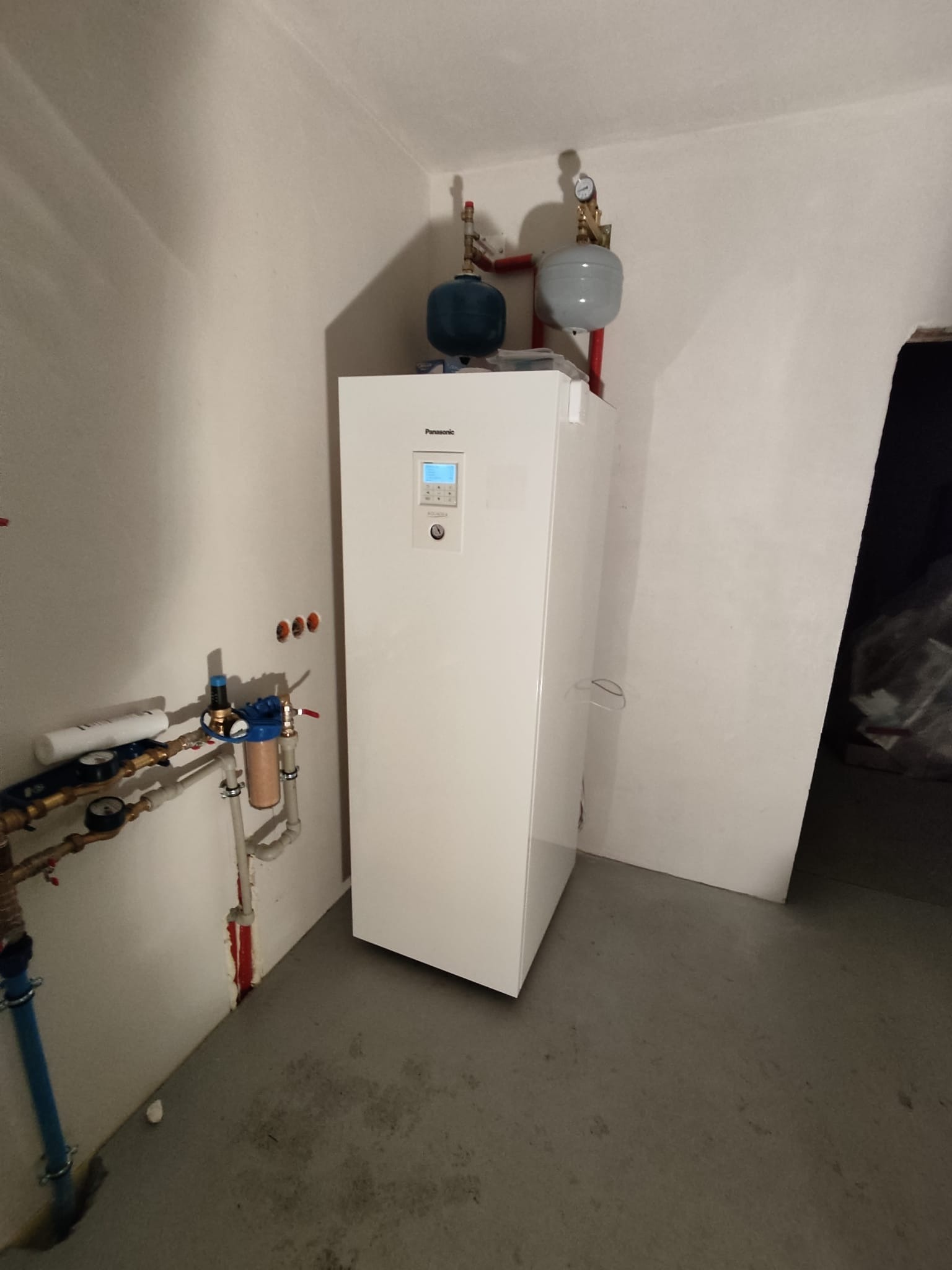 Biała jednostka wewnętrzna z wyświetlaczem, podłączona do instalacji hydraulicznej z dwoma zbiornikami wyrównawczymi na górze, zainstalowana w surowym pomieszczeniu z betonową podłogą.