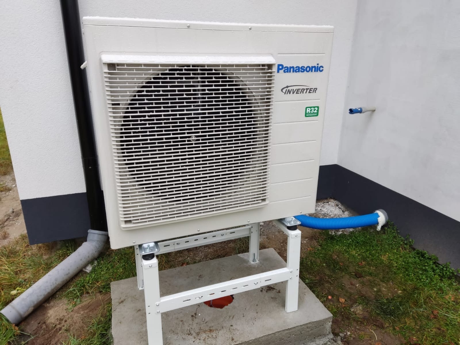 Zewnętrzna jednostka pompy ciepła Panasonic Inverter R32 na białym stelażu, zamontowana przy ścianie budynku z widoczną rurą odprowadzającą skropliny.