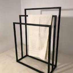 Minimalistyczny, czarny, metalowy stojak na ręczniki z trzema poziomami, z kremowym ręcznikiem na środkowym szczeblu, na tle jasnej ściany i podłogi.