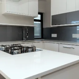 Nowoczesna kuchnia z białymi lakierowanymi frontami, szarym szkłem na ścianie i wyspą z białym blatem oraz wbudowaną płytą gazową.