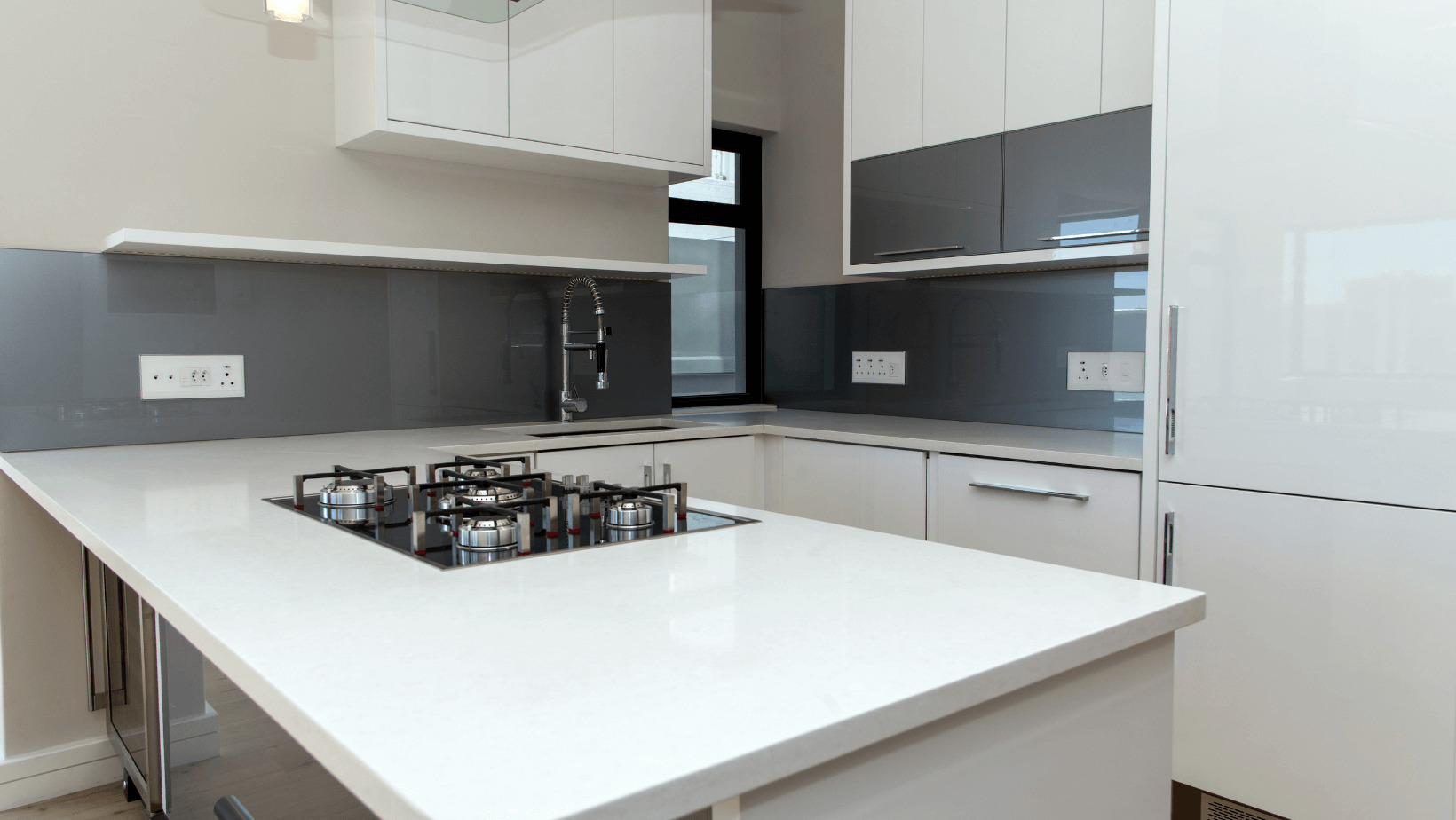 Nowoczesna kuchnia z białymi lakierowanymi frontami, szarym szkłem na ścianie i wyspą z białym blatem oraz wbudowaną płytą gazową.