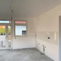 Wnętrze w trakcie remontu: białe ściany przygotowane do malowania, częściowo położone panele szklane na ścianie, surowa podłoga, widoczne instalacje elektryczne i okna z widokiem na zieleń.