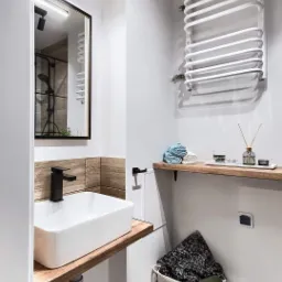 Nowoczesna łazienka po remoncie: biała umywalka nablatowa z czarną armaturą na drewnianym blacie, grzejnik drabinkowy, lustro z oświetleniem LED i dekoracyjna półka z akcesoriami.