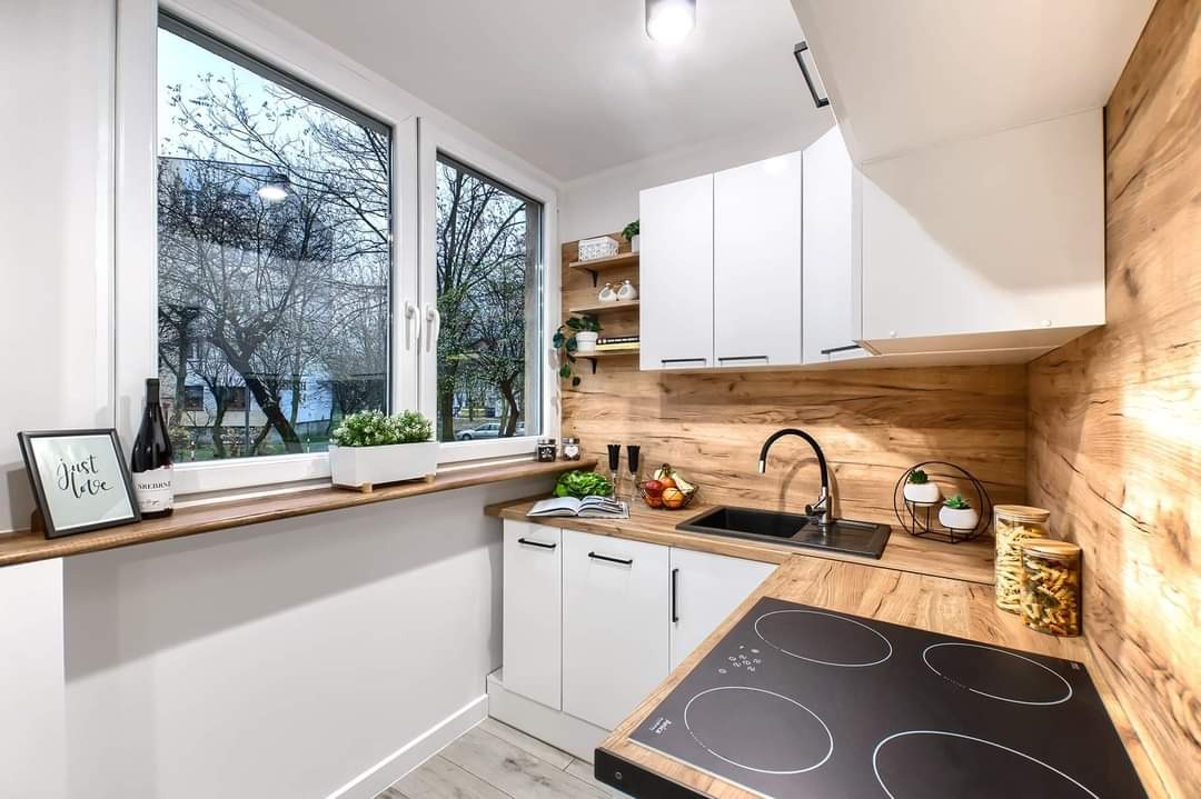 Nowoczesna kuchnia po remoncie: białe szafki z drewnianym blatem i panelem ściennym, czarny zlew i płyta indukcyjna, dekoracje okienne i półki z roślinami.