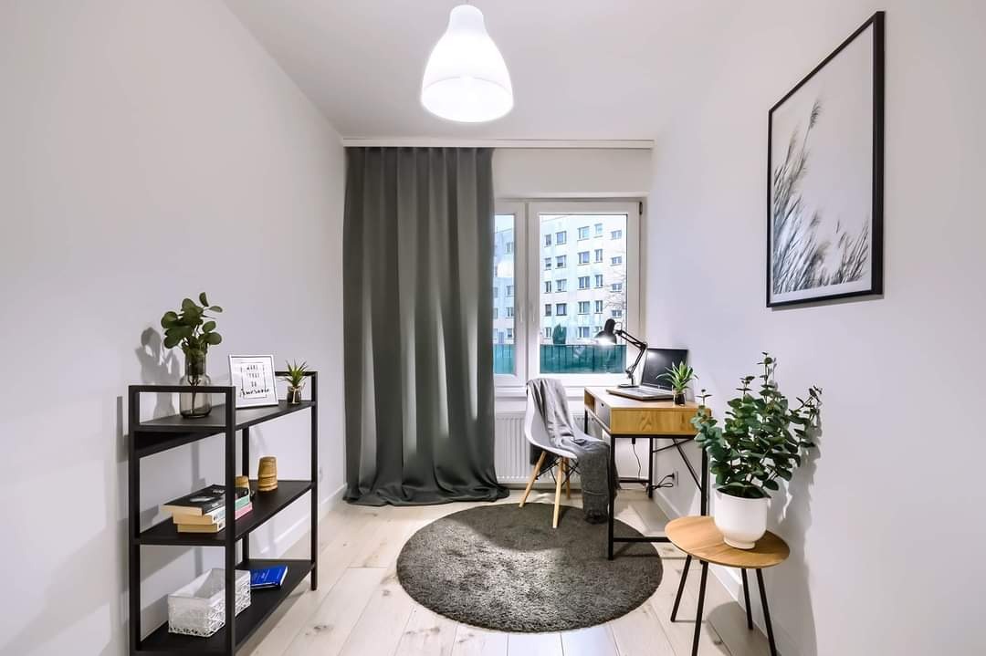 Jasne wnętrze pokoju po remoncie: biurko z laptopem, regał z książkami, okrągły dywan i szare zasłony w bloku z wielkiej płyty.