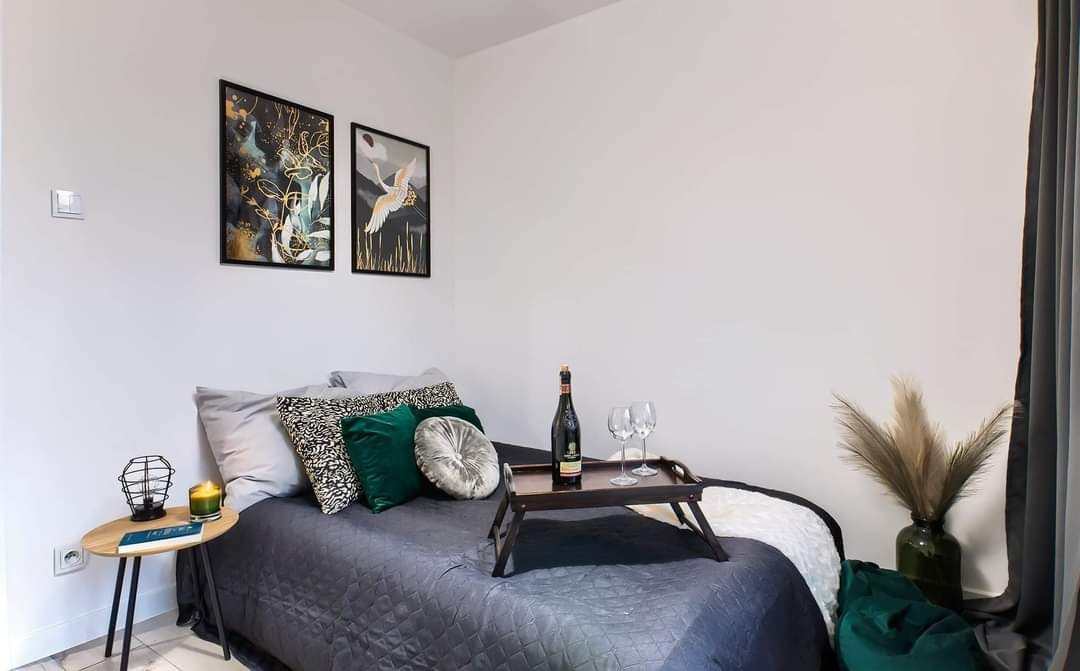 Sypialnia po remoncie: łóżko z szarą narzutą w romby, poduszki w odcieniach szarości, zieleni i czerni, stolik nocny z lampką i świecą, na łóżku taca z butelką wina i kieliszkami, na ścianie obrazy...