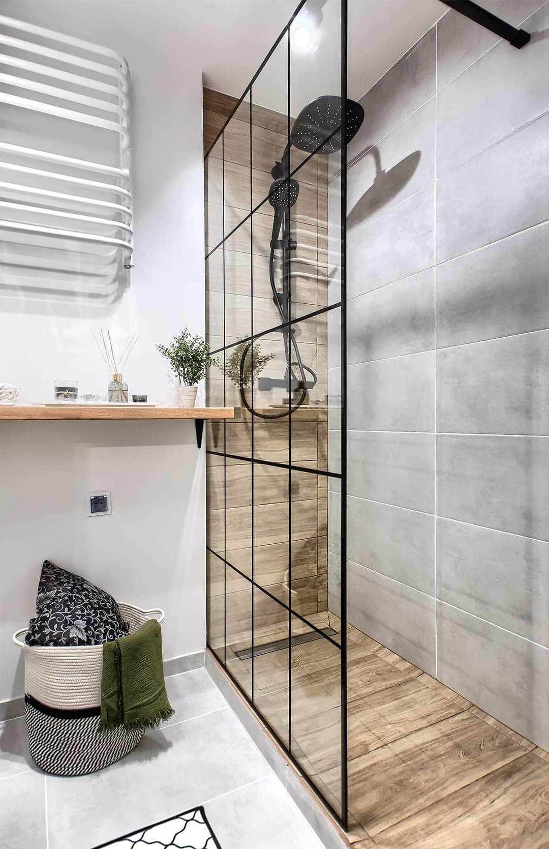 Nowoczesna łazienka z kabiną prysznicową typu walk-in z czarnymi profilami, wykończona płytkami imitującymi drewno i szare płytki, z dekoracyjnym koszem na tekstylia i grzejnikiem drabinkowym.