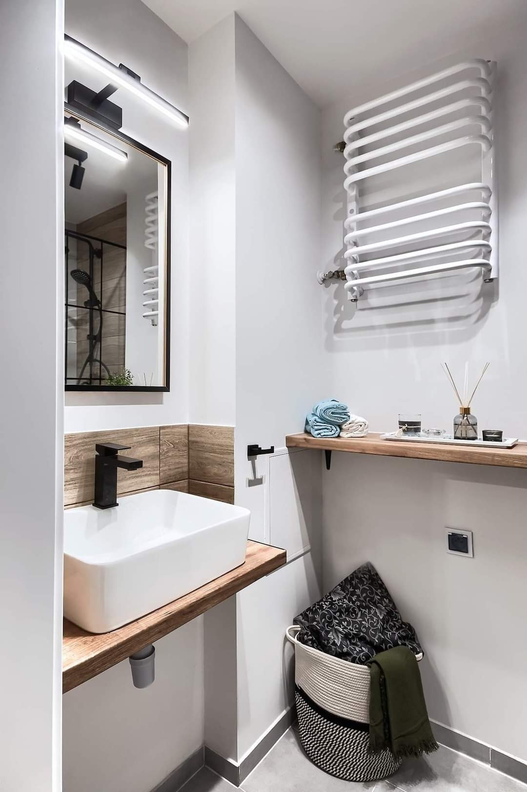 Nowoczesna łazienka po remoncie: biała umywalka nablatowa z czarną armaturą na drewnianym blacie, grzejnik drabinkowy, lustro z oświetleniem LED i dekoracyjna półka z akcesoriami.