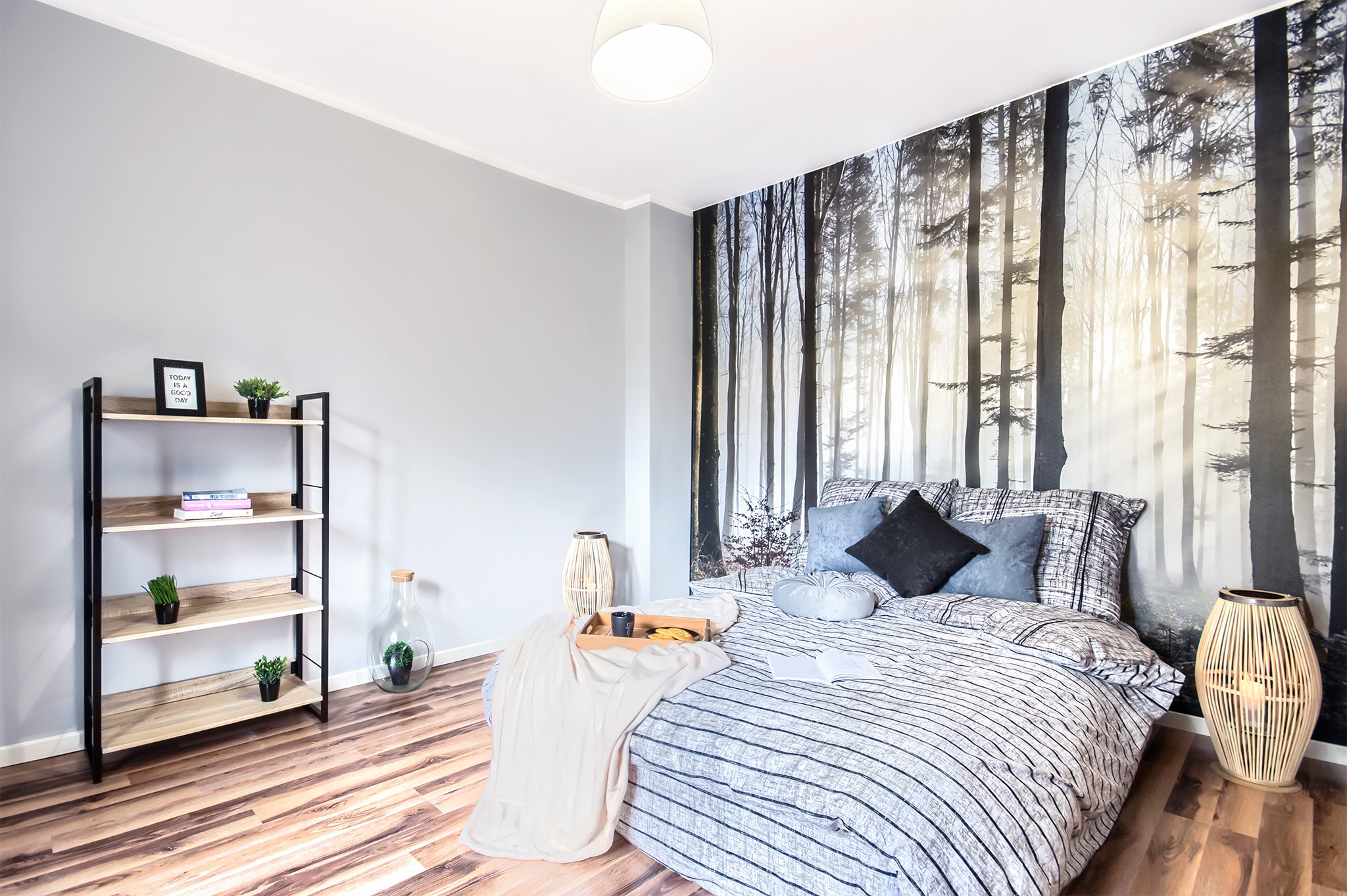 Nowoczesna sypialnia z fototapetą leśną, drewnianą podłogą, metalowym regałem z dekoracjami i łóżkiem z narzutą w paski, aranżacja po remoncie kawalerki.
