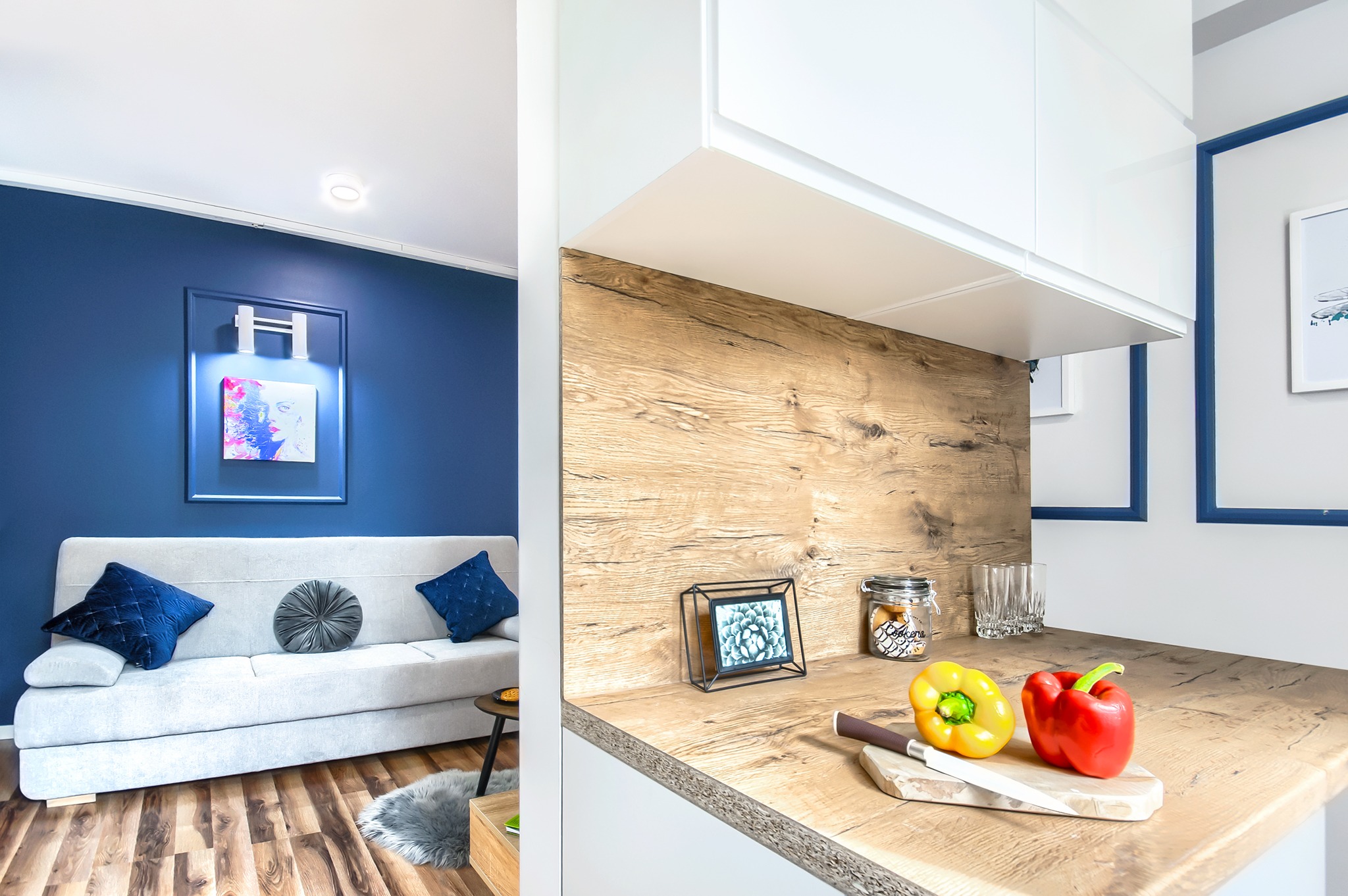 Nowoczesne wnętrze kawalerki po liftingu w Jeleniej Górze: błękitna ściana z podświetlonym obrazem nad jasnoszarą sofą z welurowymi poduszkami, drewniana podłoga i blat kuchenny z żółtą i czerwoną...